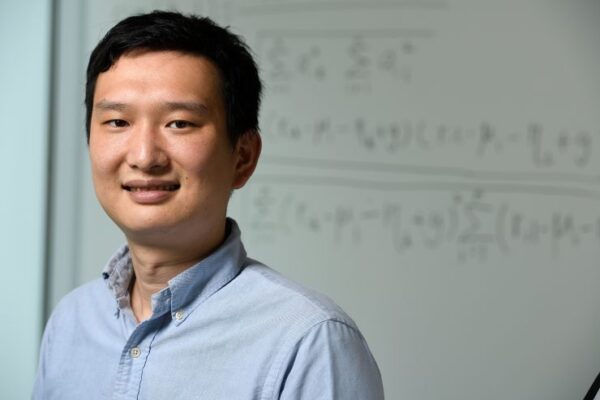 Headshot of Yinzhi smiling in a light blue button down, in front of a white board covered in an equation